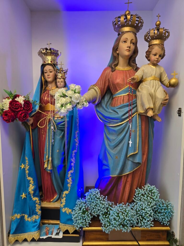 Two crowned Madonna statues displayed side by side inside a niche at the CVM Social Centre. On the left is Madonna Monserrato, dressed in blue and red robes with gold detailing, holding the Child Jesus and a bouquet of red and white flowers. On the right is Madonna Maggiore, wearing blue and rose-colored robes, holding the Child Jesus and a rosary. Both figures wear ornate gold crowns. Blue accent lighting illuminates the background, and floral arrangements are placed at the base of the statues.